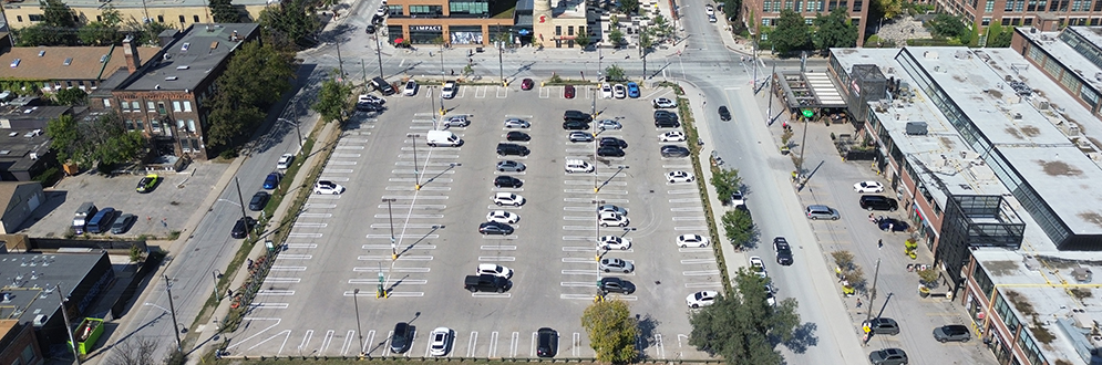• Aerial view of 34 Hanna Avenue captured by a drone, showcasing a large parking lot with numerous cars, surrounded by various buildings including multi-story office buildings and smaller structures within Liberty Village.