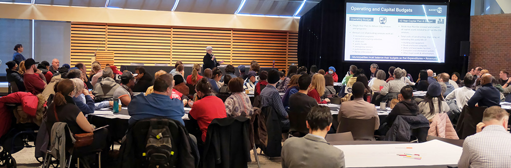 A large group of people seated around tables watching a presentation. The title on the screen at the front of the room reads Operating and Capital Budgets.