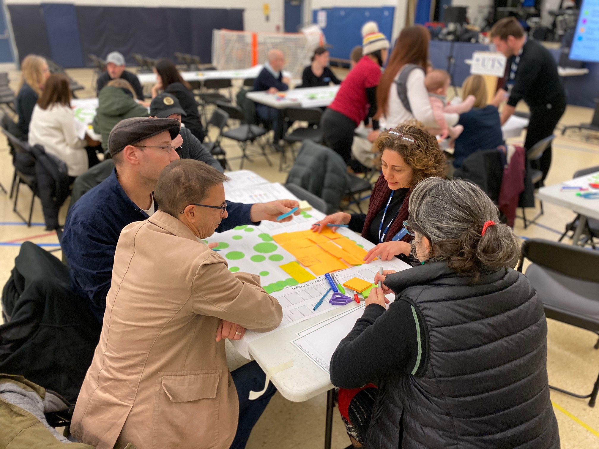 A group of workshop participants sit at a table, writing comments on sticky notes and placing them on a map of Osler Playground. Several other groups of participants are visible working at tables in the background.