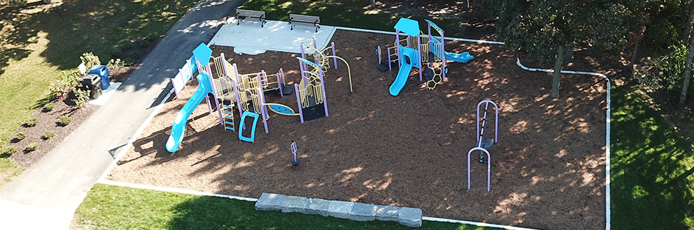 Aerial view showing compeletd playground. New playground shows a senior playstrucure, junior play structure, one swing set, one springer, and one accessible play panel. There are two benches in a concrete seating area at the south-east corner and a row of armour stone seating on the northern edge of the playground.