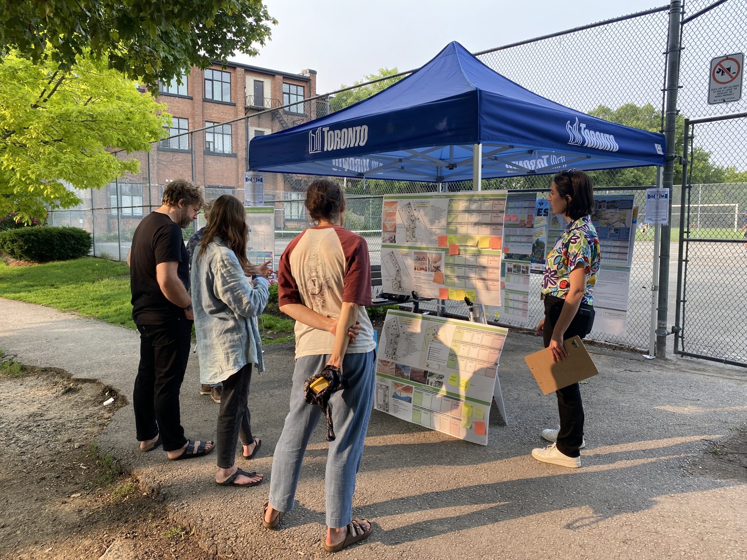 Community members stand in front of a pop-up tent, studying two display boards that present the preferred design for the community recreation centre along with feedback written on sticky notes. More display boards are visible at the back of the tent.