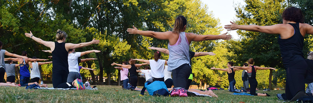 People of mixed ages and ethnicities participating in an outdoor fitness class on the grass in a park; arms stretched wide.