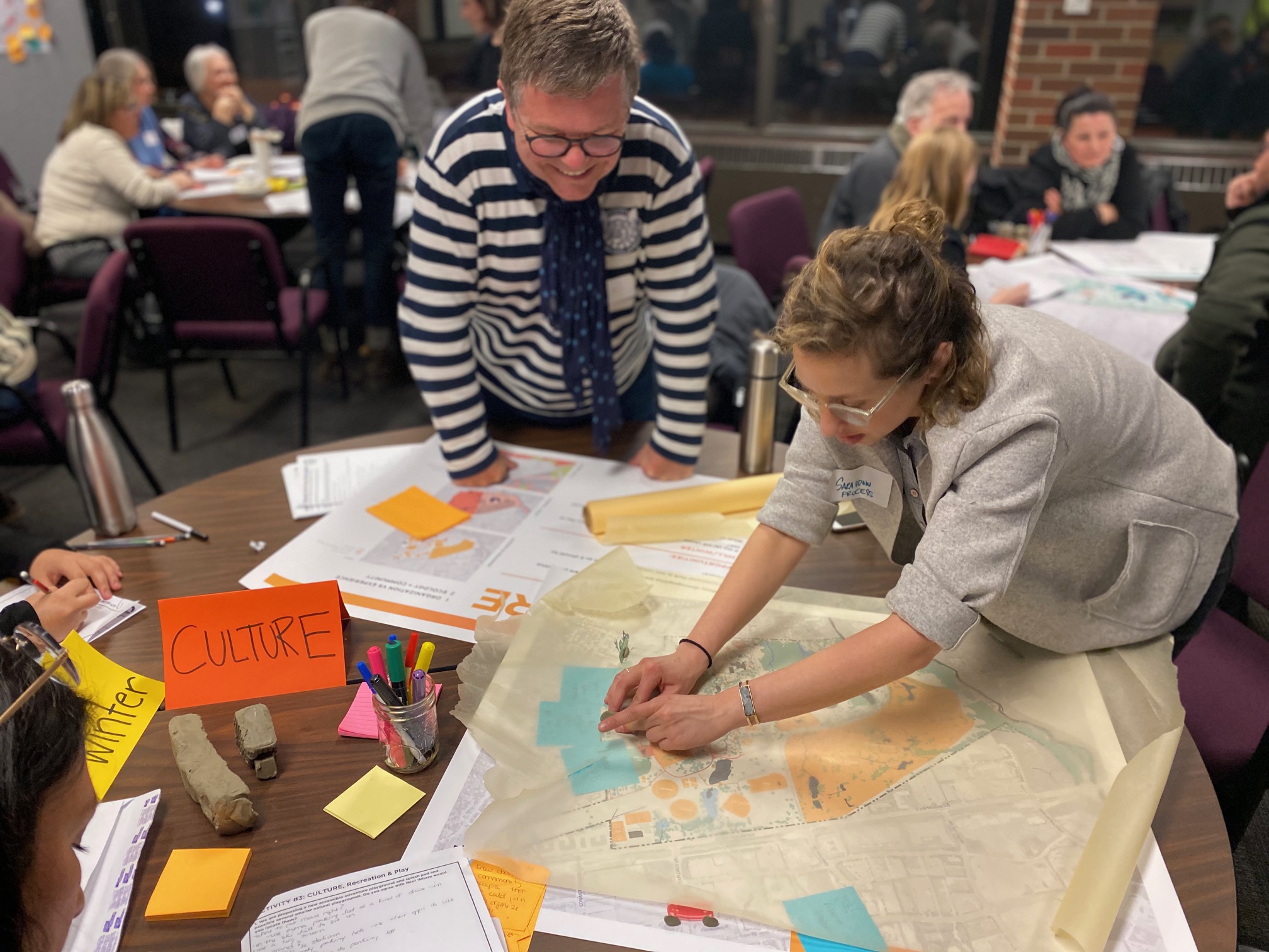 Two workshop participants engage with a map of the park spread across a table, surrounded by an orange sign labelled ‘CULTURE,’ sticky notes, markers and printed activity sheets. Other participants are visible discussing at tables in the background.