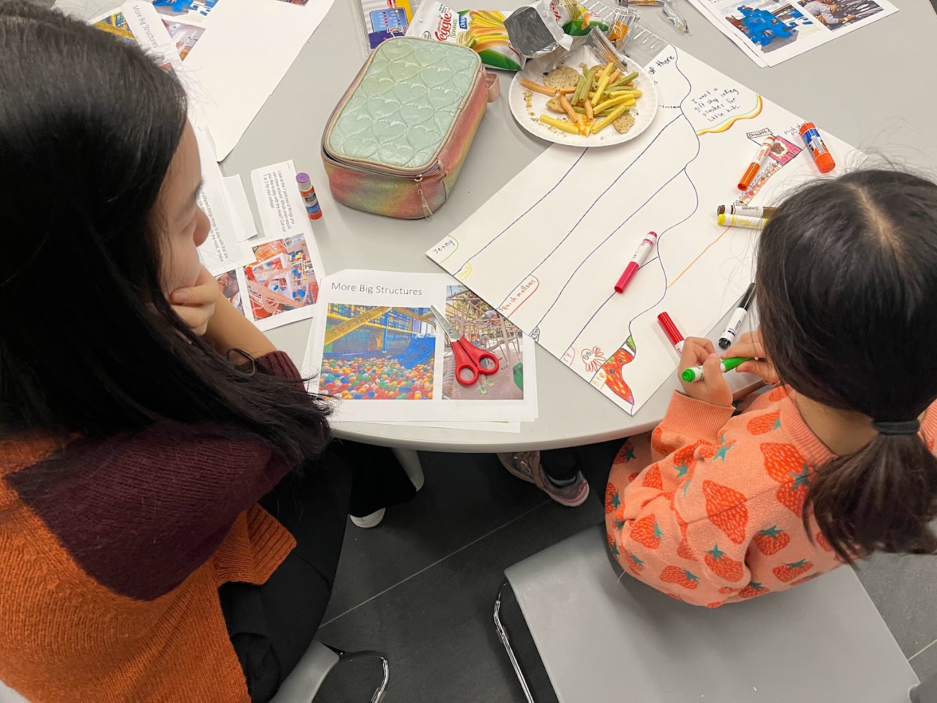 A child sits at a table, using markers to draw and write ideas for the indoor play space on a sheet of paper, as a workshop facilitator looks on. On the table are scissors, glue sticks, printed images of indoor play structures and snacks.