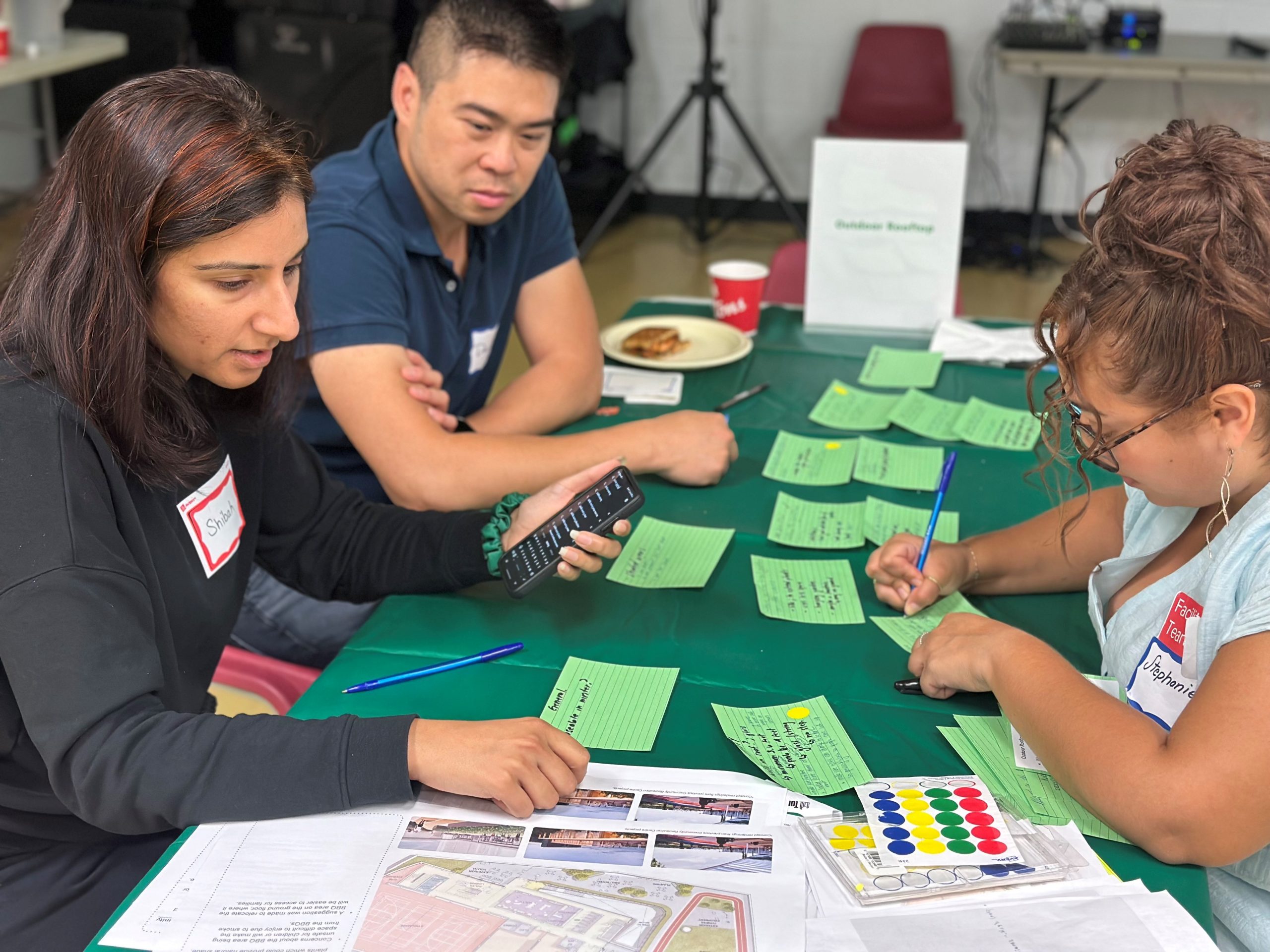 Two participants look at designs for the community recreation centre on one side of a table while a workshop facilitator writes notes on a green index card. Other green index cards with written feedback and dot stickers are displayed on the table.