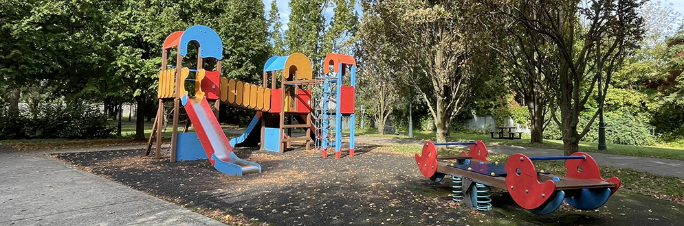 A playground in the fall showing a large play structure in orange, blue and red, with two slides, and a standalone see-saw.