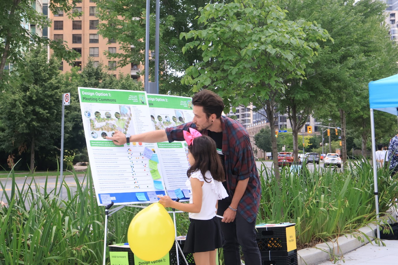 A young community member reviews two display boards, each presenting a different design option for the park, beside a City staff member. The display boards are covered with small dot stickers and sticky notes with written comments.
