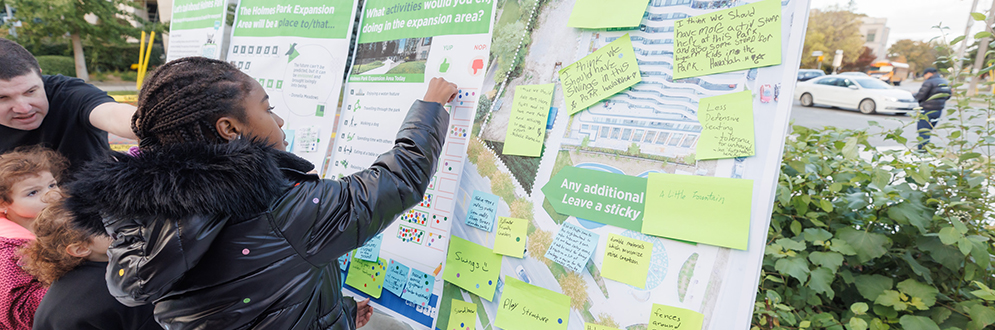 Four display boards with project information at a pop-up event. A child is placing sticky dots to share their preferences. The display board in the foreground has post-it notes with comments.