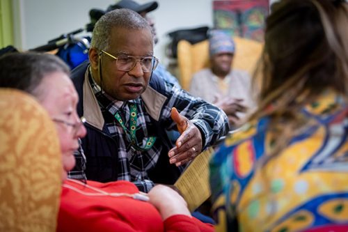 People sit together in a small group having a discussion, with one person gesturing while speaking during a community meeting.