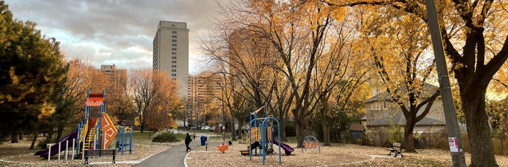 Photograph with a view of Benner Park, looking south during the fall season towards a playground with trees, open space, and people using a park path. Single detached dwellings are visible on the right side of the image, with other single detached and high rise buildings adjacent near Ridelle Avenue visible in the background.