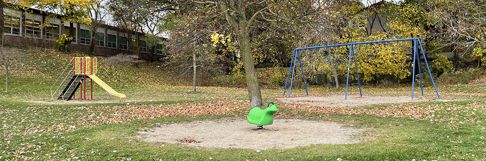 Park Lawn Park playground surrounded by open lawn and mature trees. The playground is located next to a school at the base of a hill. A home can be seen in the background.