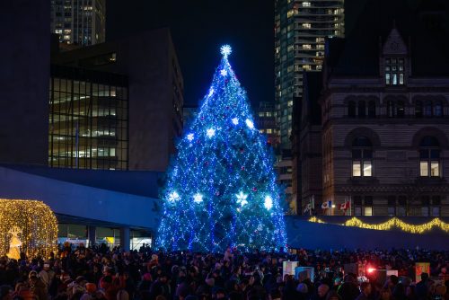 A large Christmas tree adorned with sparkling lights and ornaments in Nathan Phillips Square, illuminated against the night sky with festive decorations.