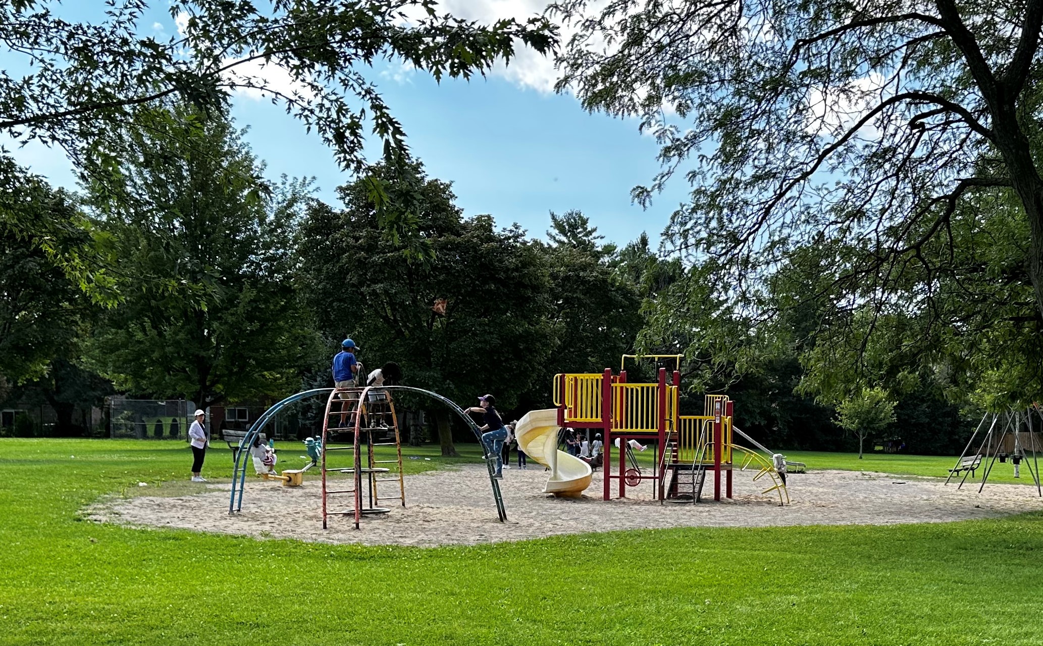 Image of the playground before construction, featuring a sand surface, one climbing structure, a junior play structure in the foreground, and two swing sets and a seesaw in the background.