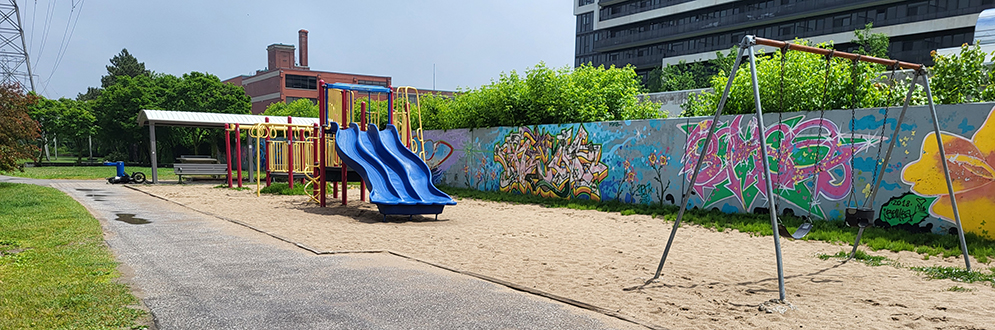 Playground area with a blue slide, swings, and sand, surrounded by a paved path. A mural with colorful graffiti decorates the wall in the background, with greenery and buildings visible beyond the park.
