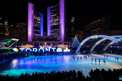 People ice skating in Nathan Phillips Square, with holiday lights surrounding the rink and Toronto's city skyline in the background.