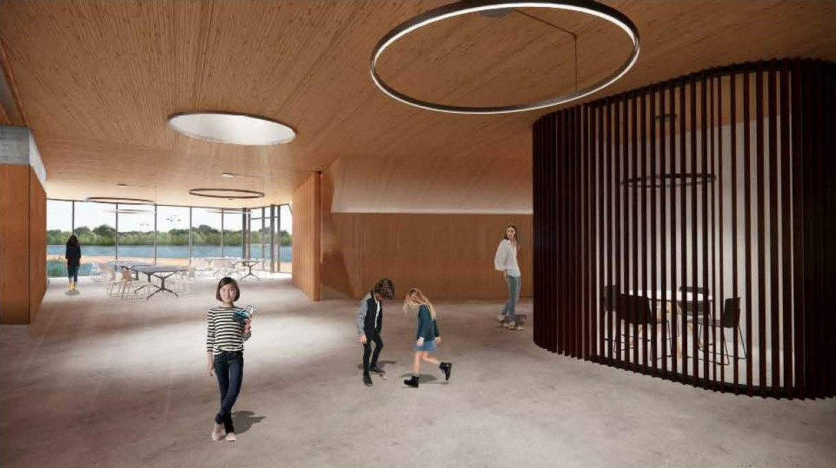 Rendering of an area within the recreation building. The spacious interior has concrete floors, wood finishes on the walls and ceiling, and large circular lights hanging from the ceiling. On the right, a curved partition made of long, vertical wooden strips creates a division for the staff meeting room, which has a small table and chairs. In the background, additional tables and chairs are visible surrounded by floor-to-ceiling windows that look out to a terrace, pond and trees.
