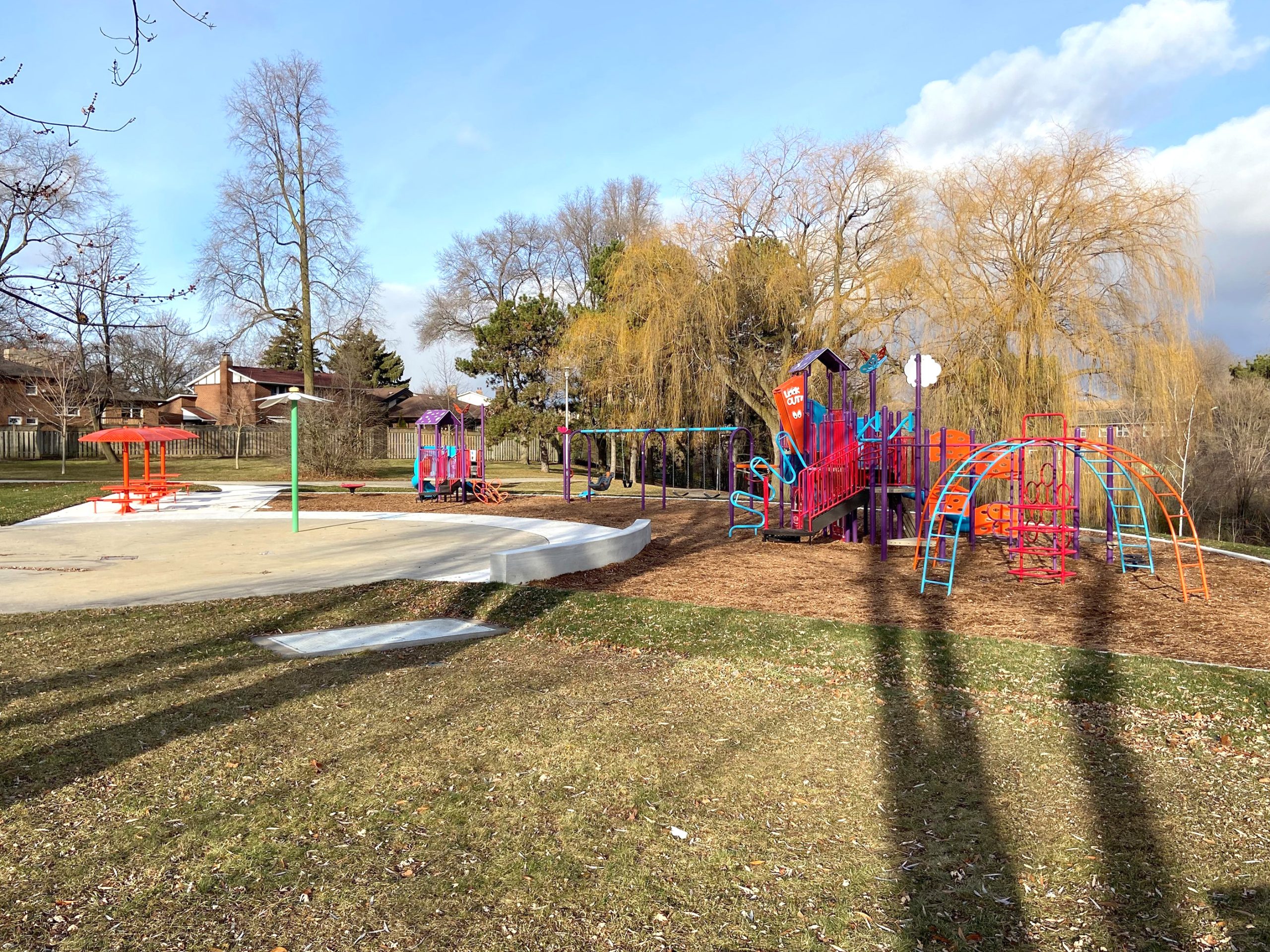 The new Stephen Leacock Park playground looking north-west from the existing lawn area. The playground has a splash pad and open lawn in the west foreground and is surrounded by a walkway, mature willow trees and residences in the background.