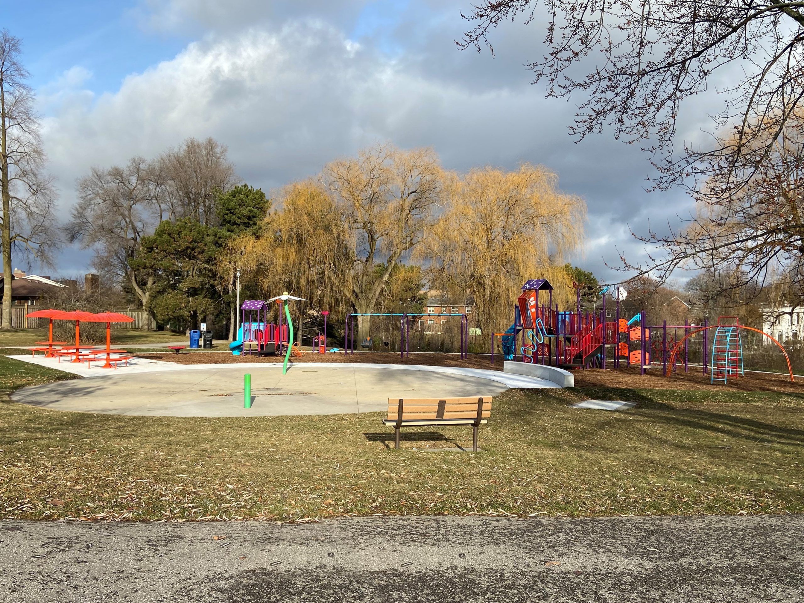 The new Stephen Leacock Park playground looking north from the existing walkway. The playground has a splash pad and open lawn area with benches in the foreground and is surrounded by mature willow trees and residences in the background.