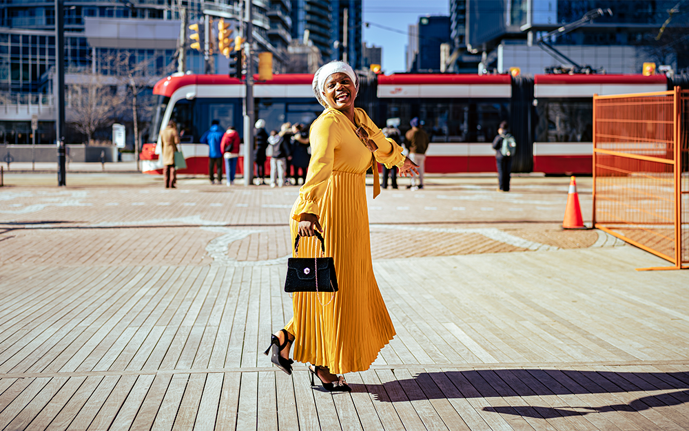 A black woman in a yellow dress and white turban smiling at the camera standing in front of a street car