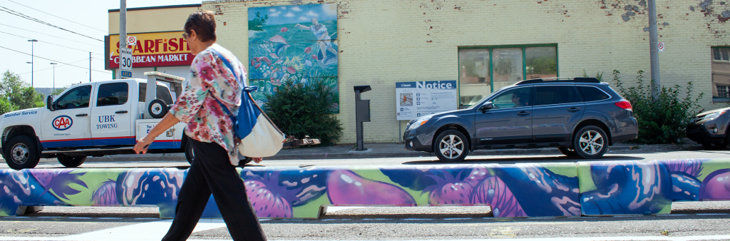 A person walks on a section of a roadway that has been allocated to pedestrian use. Colourful concrete barriers separate the pedestrian space from the motor vehicle travel lanes.