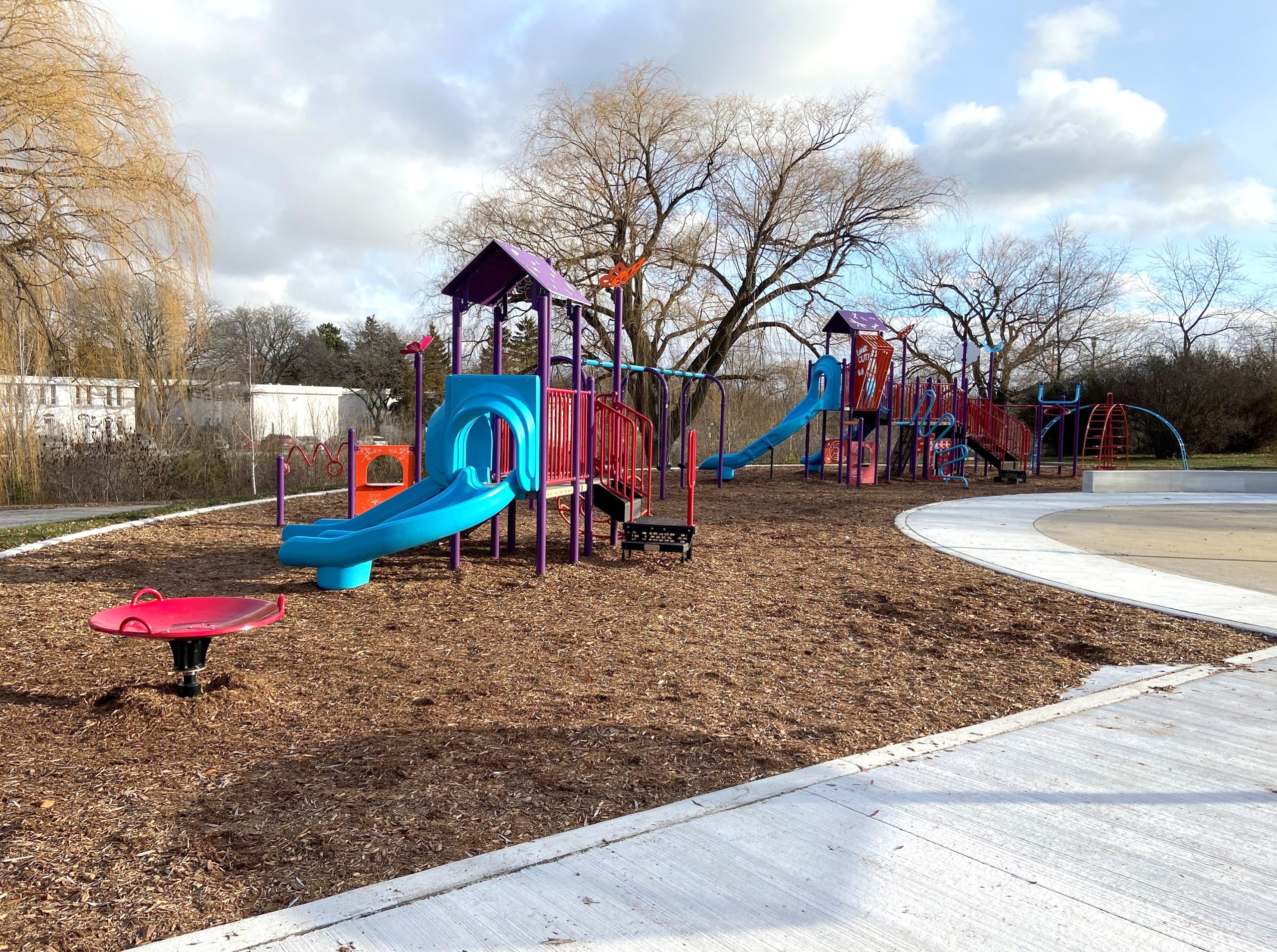 The new Stephen Leacock Park playground looking east from the new walkway. The playground is surrounded by a walkway, mature trees and residences in the background.