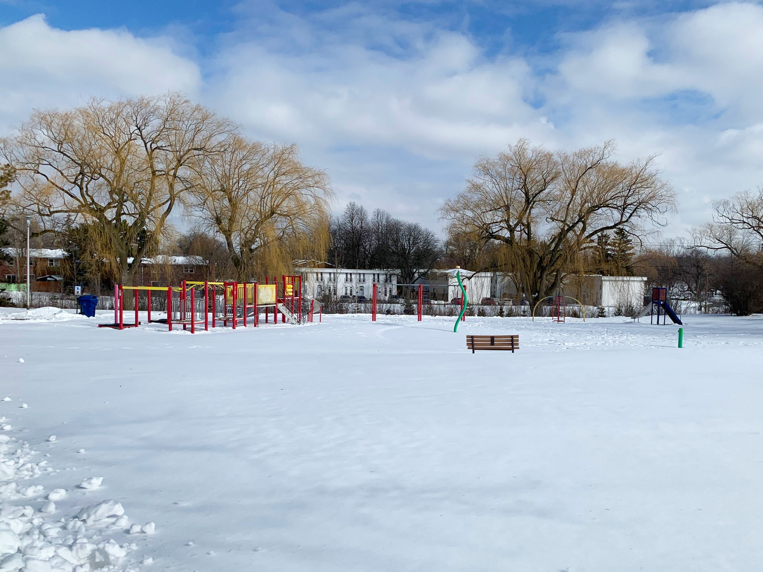 Stephen Leacock Park playground before construction looking north-east from the existing walkway. The playground is surrounded by mature willow trees and residences in the background.