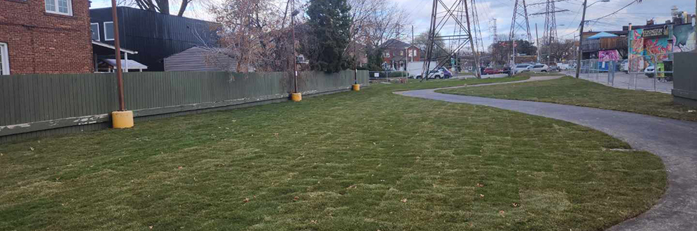 Open grassy area with newly installed sod, curved paved pathway, utility poles, and adjacent residential fencing on one side. Overhead power lines and murals on nearby buildings are visible in the background, under a partly cloudy sky.
