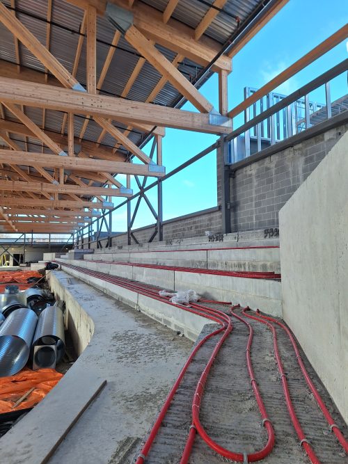 The interior of the arena under construction, focusing on the tiered concrete bleachers with exposed heating pipes. Construction materials, including ducts and tarps, are piled at the foot of the bleachers. The roof is supported by wooden trusses with metal decking. Exterior walls made of concrete blocks and steel framing are in progress. 