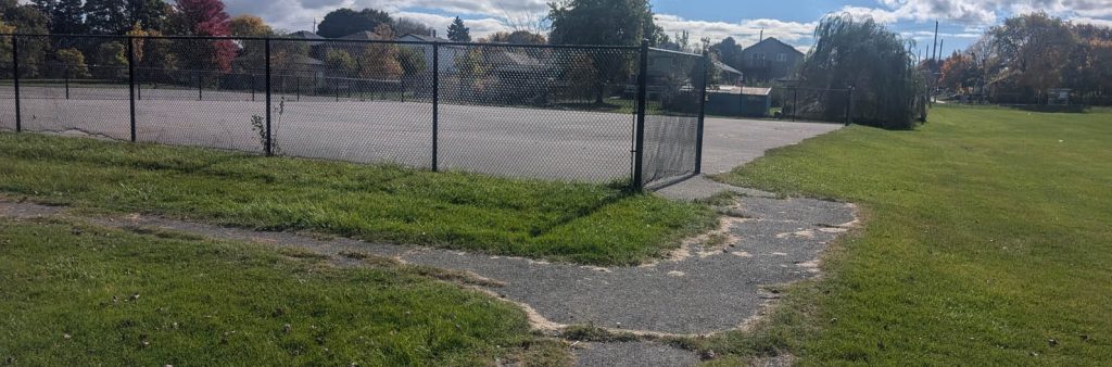A grassy park area with a worn paved path running along and leading to a large, fenced sports court. More grassed open space is visible in the background, in addition to trees and houses under a partly cloudy sky.