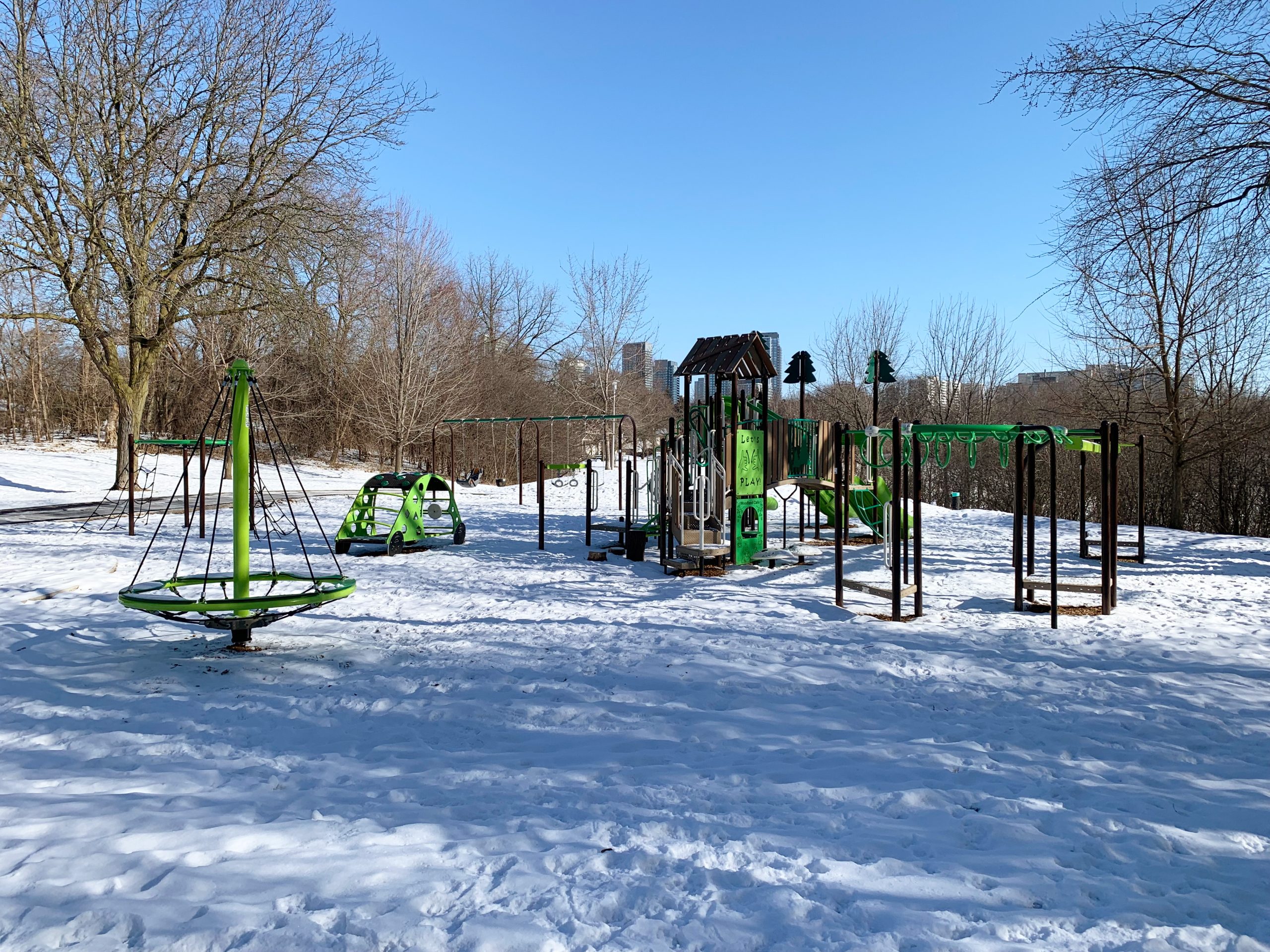 The new Havenbrook playground looking east from the tennis club building. The playground and park are snow covered with the main walkway visible in the background to the left and dense vegetation to the right.