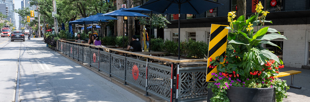 A restaurant patio situated in the curb lane of the street with blue umbrellas and a big planter.