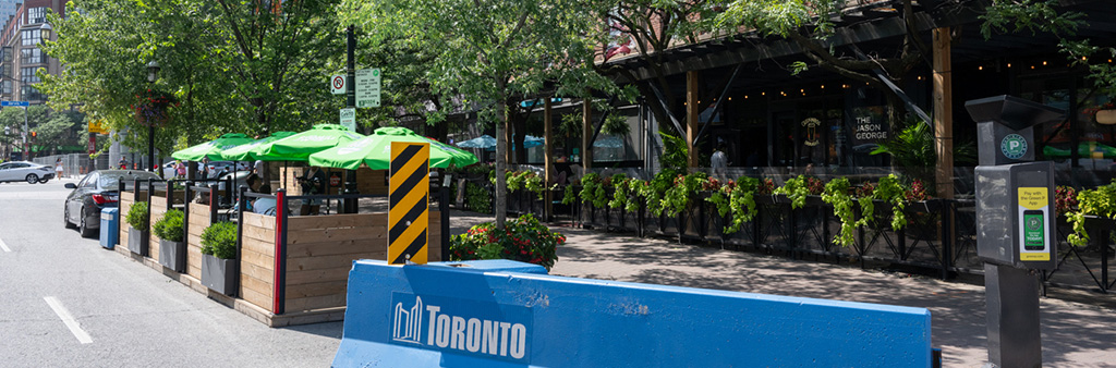 A restaurant patio with green umbrellas situated in the curb lane of the street with lots of greenery and a large blue painted concrete barrier with a white City of Toronto logo painted on it.