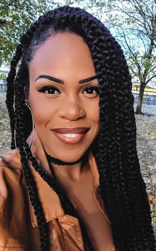 A selfie portrait of Kanisha Dabreo, a Black woman with long braided hair, smiling. She is outdoors, wearing a brown jacket, with trees in the background.
