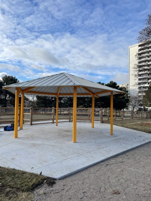 An open-sided shade structure with a grey roof supported by yellow poles stands on a concrete pad atop grass and dirt. Behind the structure, a fenced open area is visible. In the background are residential buildings and trees under a partly cloudy sky. 