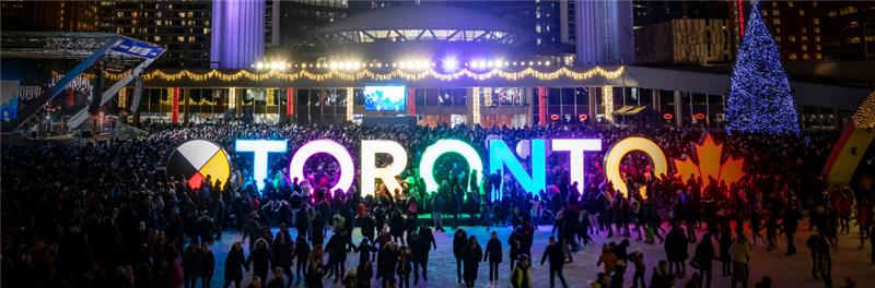 Night scene of skaters on rink in front of colourful Toronto sign, Christmas tree and City Hall.