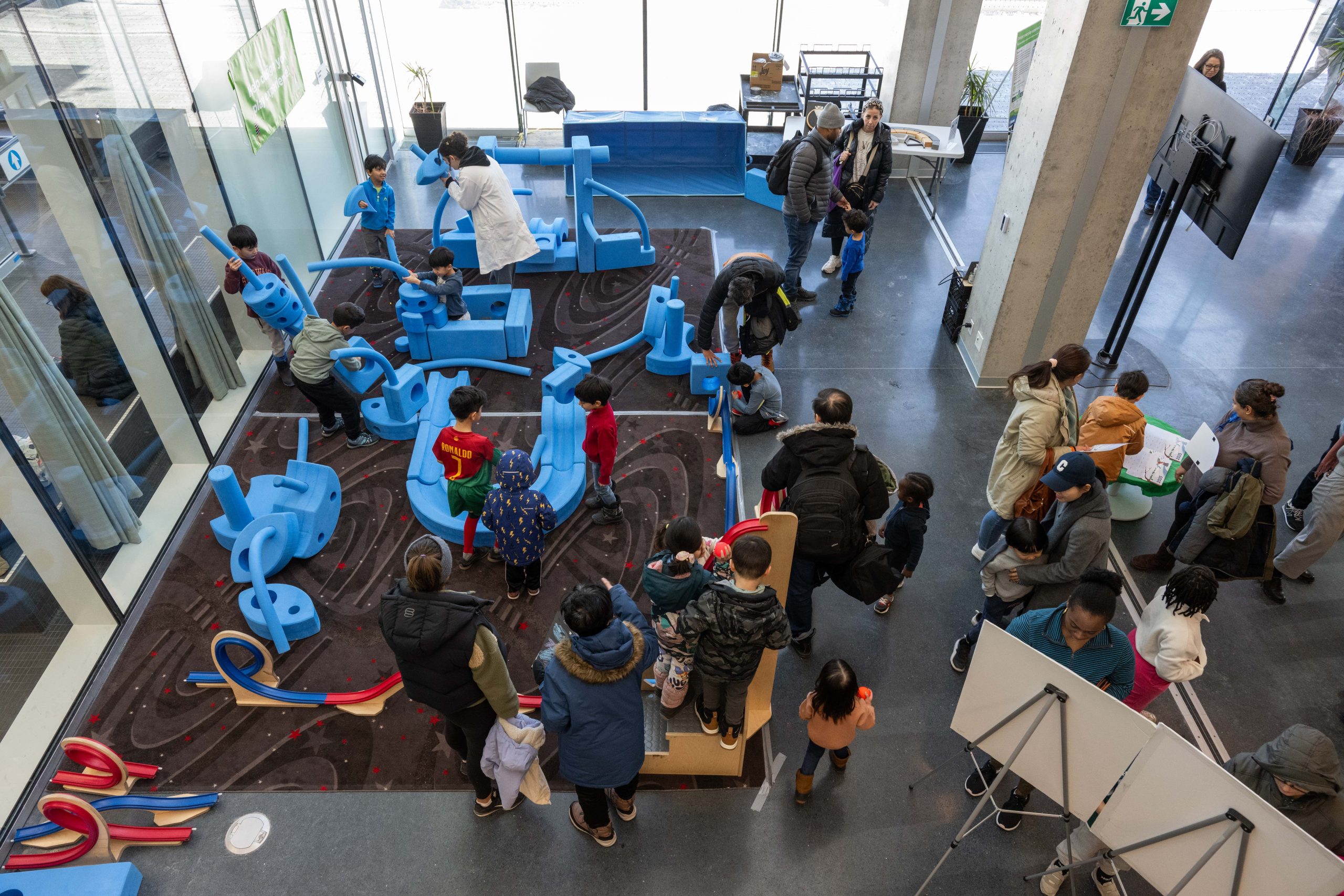 Children and adults interacting with large blue foam building blocks in a busy indoor play area. The scene is lively with various play structures and natural light from large windows.