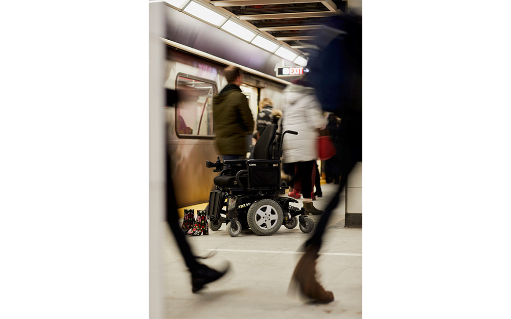 Empty electric wheelchair and decorative pair of boots sit on a subway platform with blurry people walking by.