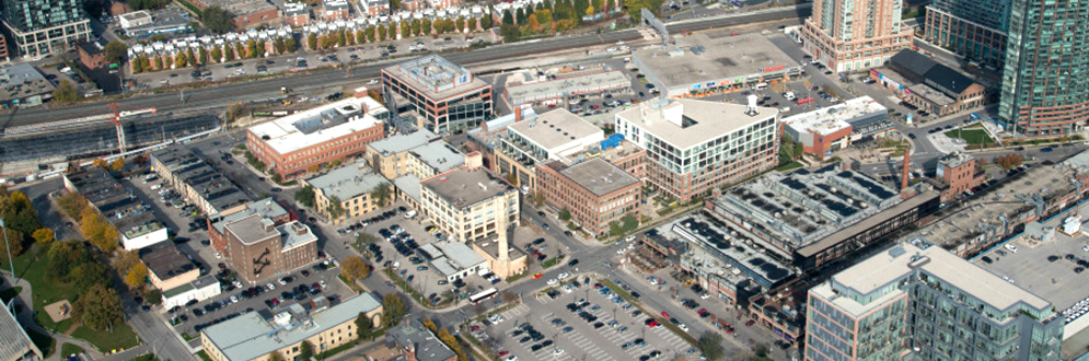 Aerial view of Liberty Village in Toronto, showcasing a mix of historic brick buildings, modern glass structures, and surface parking lots. A railway line is visible at the top of the image, running parallel to a row of townhouses and trees.