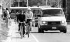 Two men biking in a bike lane on a road with a white van driving on the road next to it.