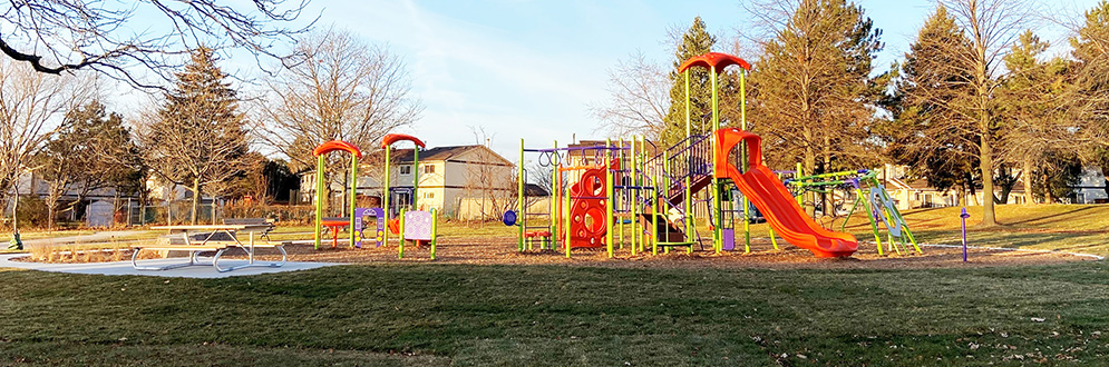 A vibrant playground with orange and green climbing structures, slides, and swing sets on a mulch play surface. Nearby, benches are placed along a paved pathway surrounded by landscaped greenery. In the background, residential houses and high-rise buildings are visible, framed by leafless trees on a clear day.