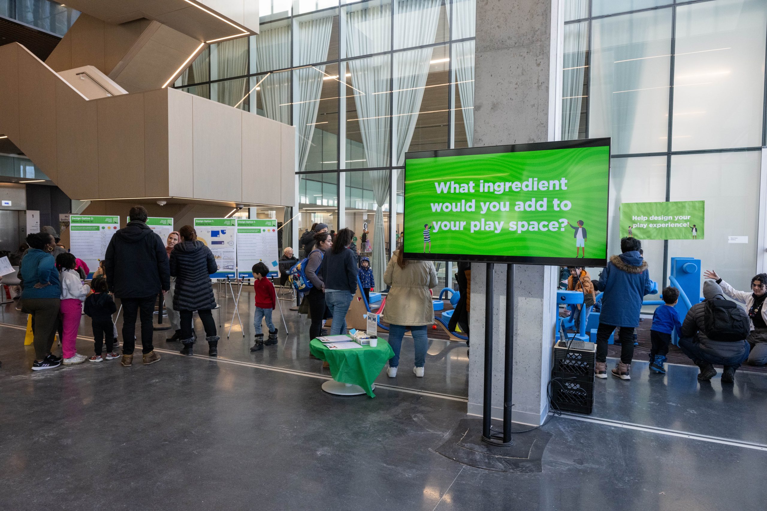 An event showcasing design options for an indoor play space. Adults and children interact with displays and activities, with boards showing play space design ideas.