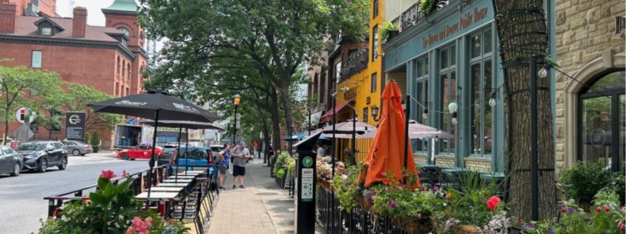 The picture shows view of existing sidewalk along Elm Street with low-rise building frontages to the right, seating patios on both sides of the sidewalk, mature trees with green leaves, planters and women and man standing on the sidewalk in the distance.