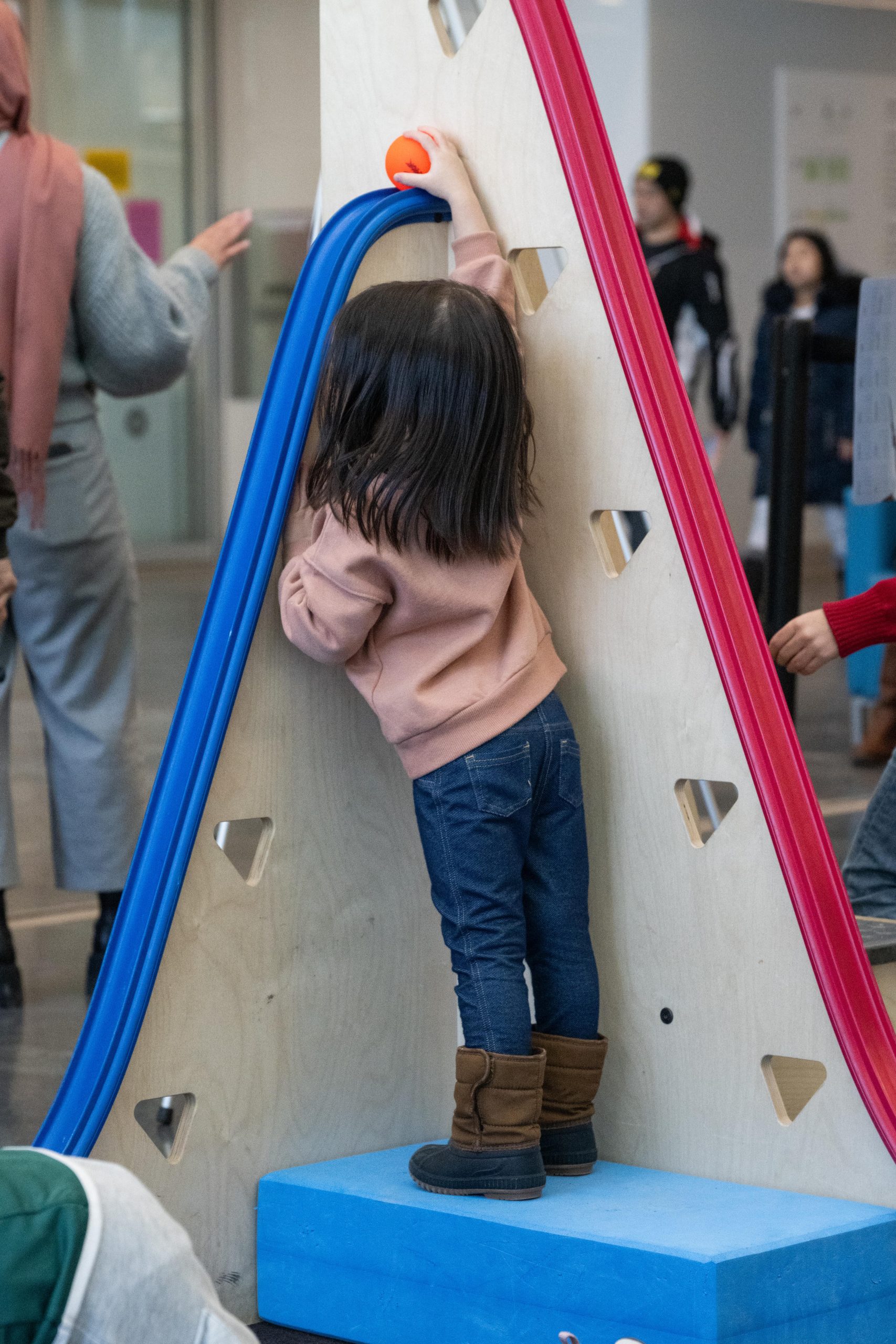A child standing on a wooden base, reaching to place an orange ball at the top of the blue roller coaster track.