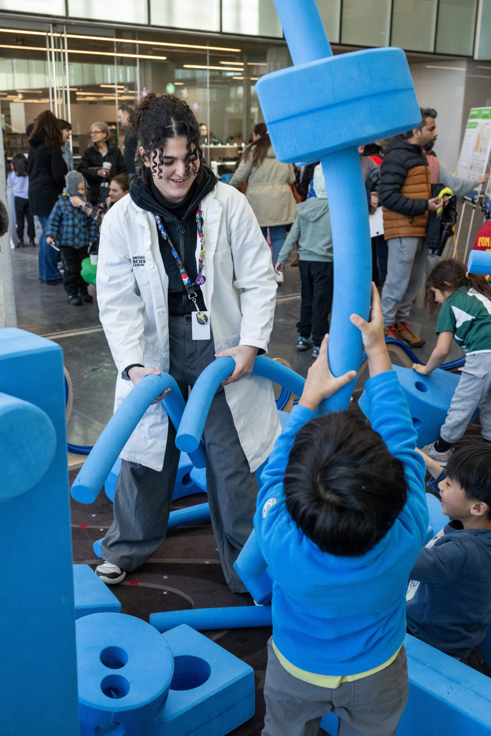 A host from the Ontario Science Centre, dressed in a white lab coat, demonstrates creative ways to play with large blue foam blocks to children at an open house event.