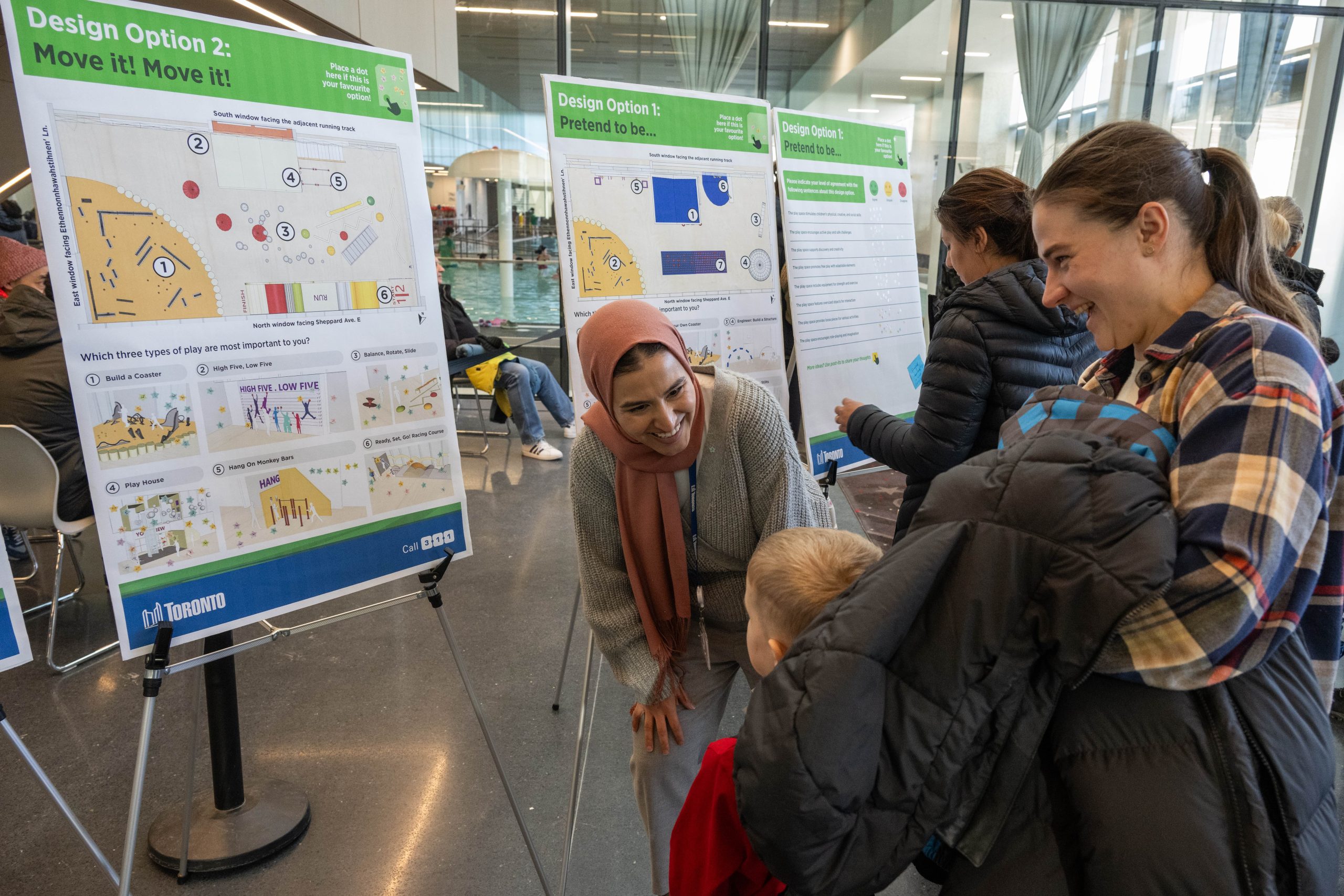Participants at an open house event examining three large boards on easels. The boards showcase different design options for an indoor play area, titled 