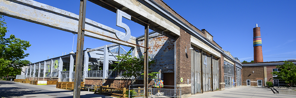 A historic brick industrial building with large metal-framed windows and remnants of an overhead structure labeled with the number '5.' The building, dated '1921,' has a partially open section revealing a modern glass structure inside. A tall smokestack with colorful bands stands in the background against a bright blue sky. The surrounding area includes paved walkways, trees, and benches.