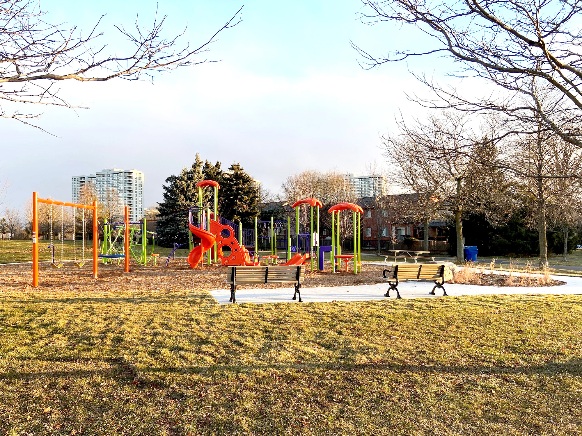 A playground in a park with colorful equipment, including swings, climbing structures, and slides in orange, green, and purple. The playground is surrounded by grass, benches, and leafless trees. In the background, there are residential buildings and a clear sky.