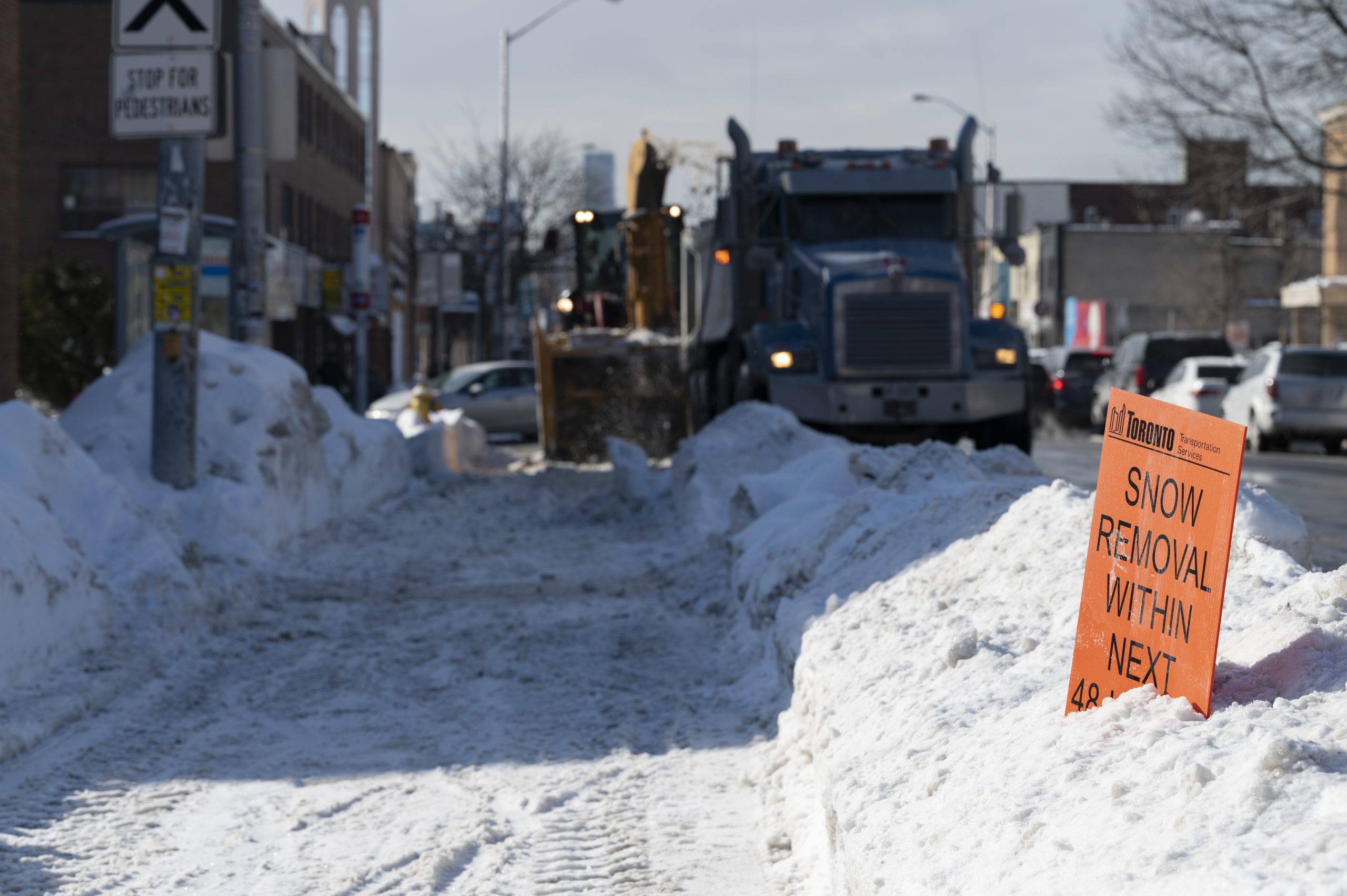 Sidewalk cleared with a snow removal sign in the snow bank and blower putting snow into dump truck in the background