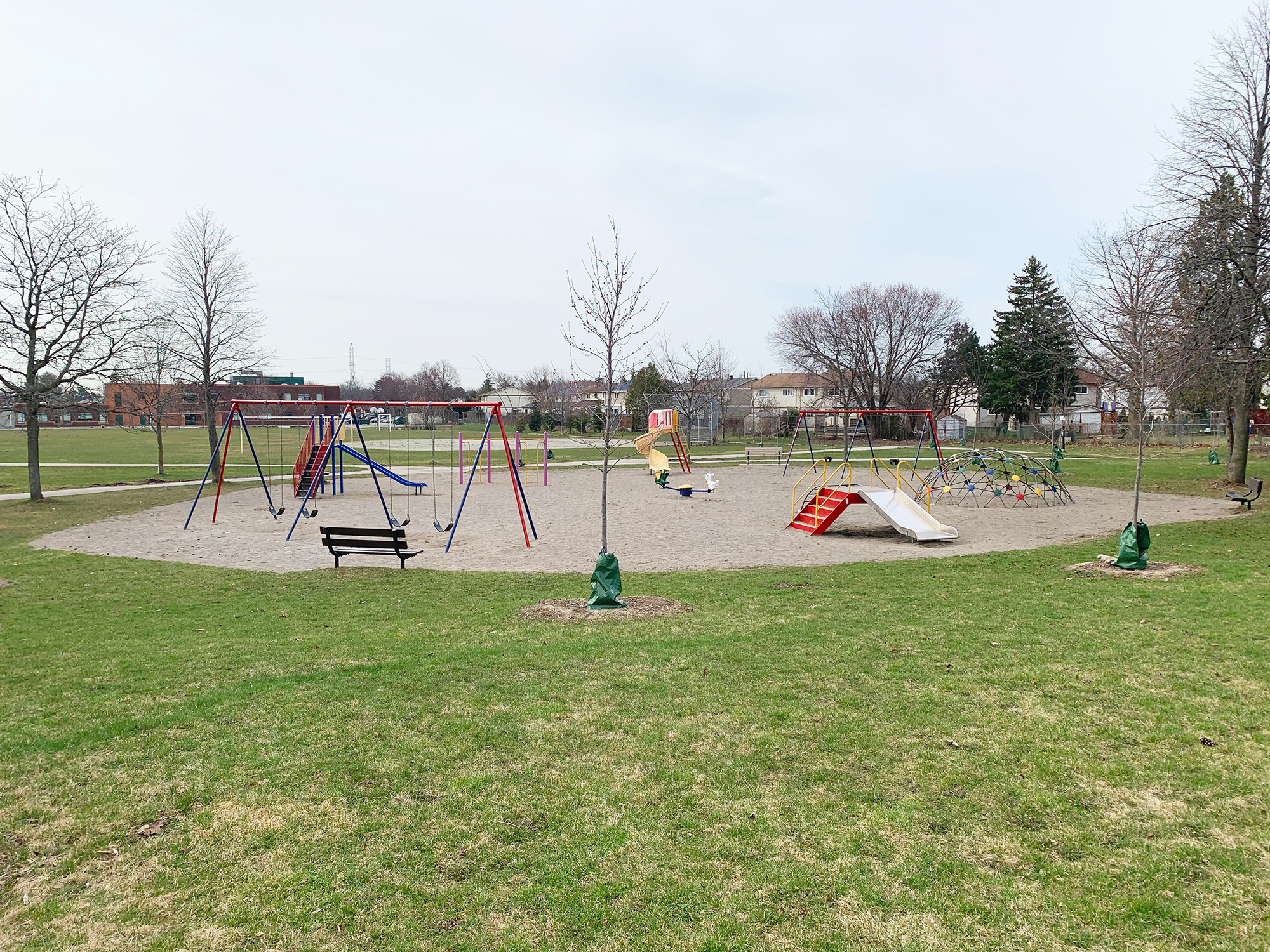 A playground with multiple swing sets, slides, and climbing structures on a sandy play area surrounded by green grass and young trees. A bench faces the playground, and residential houses are visible in the background.