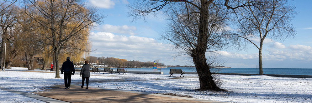 Two people walking along a boardwalk on a bright winter day, surrounded by bare trees and scenic views of a calm, expansive lake under a vibrant blue sky.